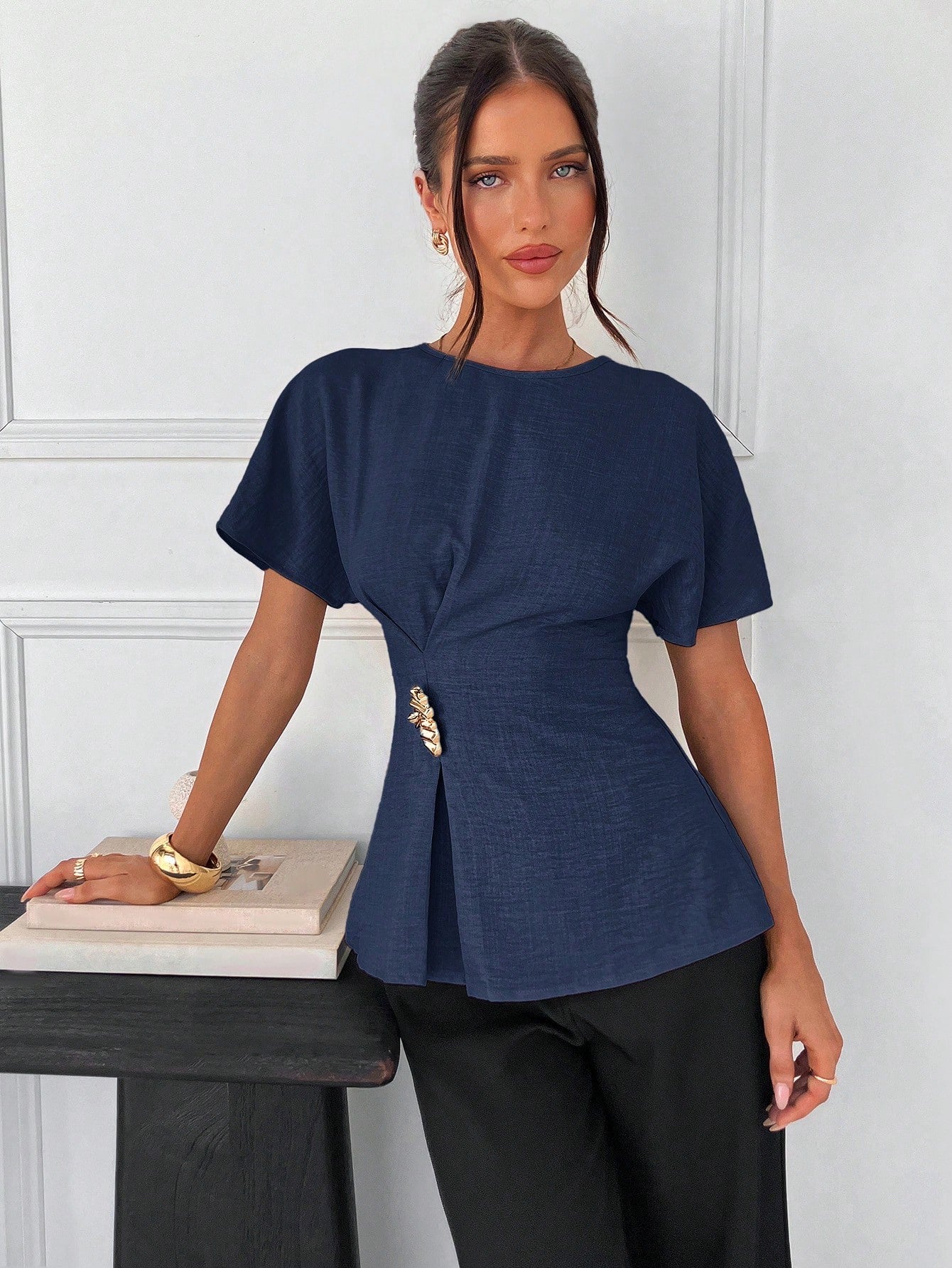 Women's navy blue short-sleeve round neck blouse with a statement gold side waist buckle paired with black trousers for an office look.