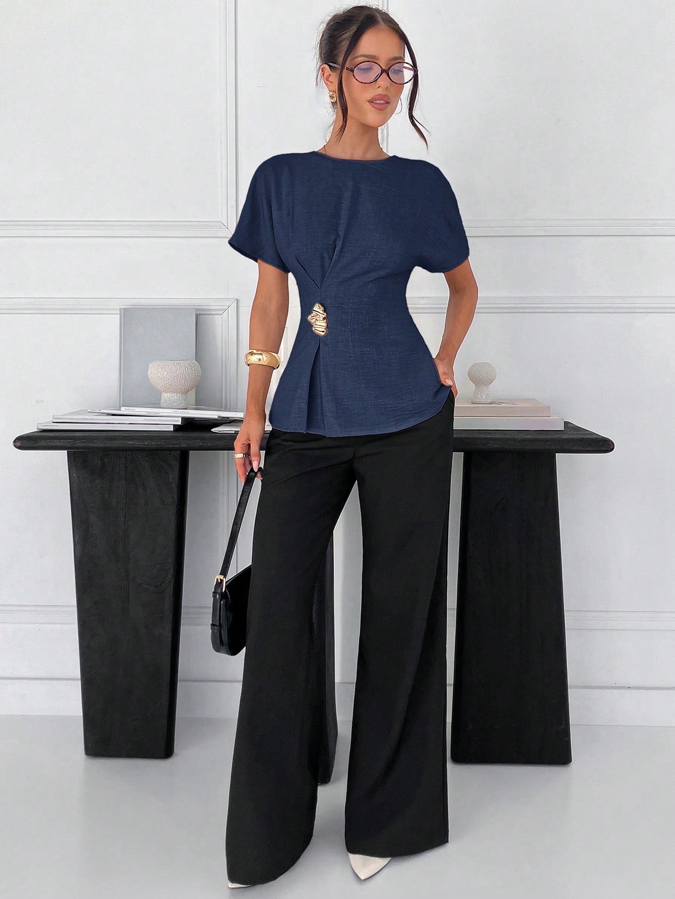 Women's navy blue short-sleeve round neck blouse with a statement gold side waist buckle paired with black trousers for an office look.