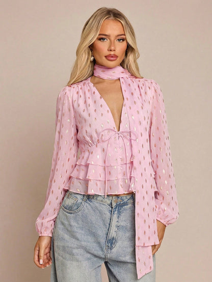 Women's pink long-sleeve chiffon blouse with metallic foil dots, a V-neck, a scarf tie, and a tiered ruffled hem paired with blue jeans.
