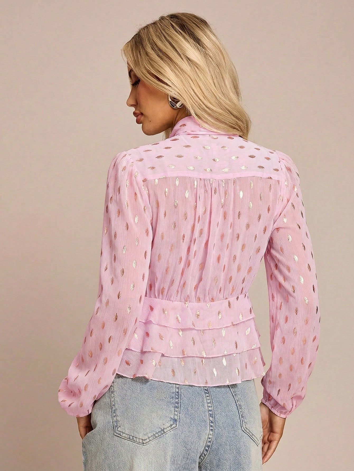 Women's pink long-sleeve chiffon blouse with metallic foil dots, a V-neck, a scarf tie, and a tiered ruffled hem paired with blue jeans.