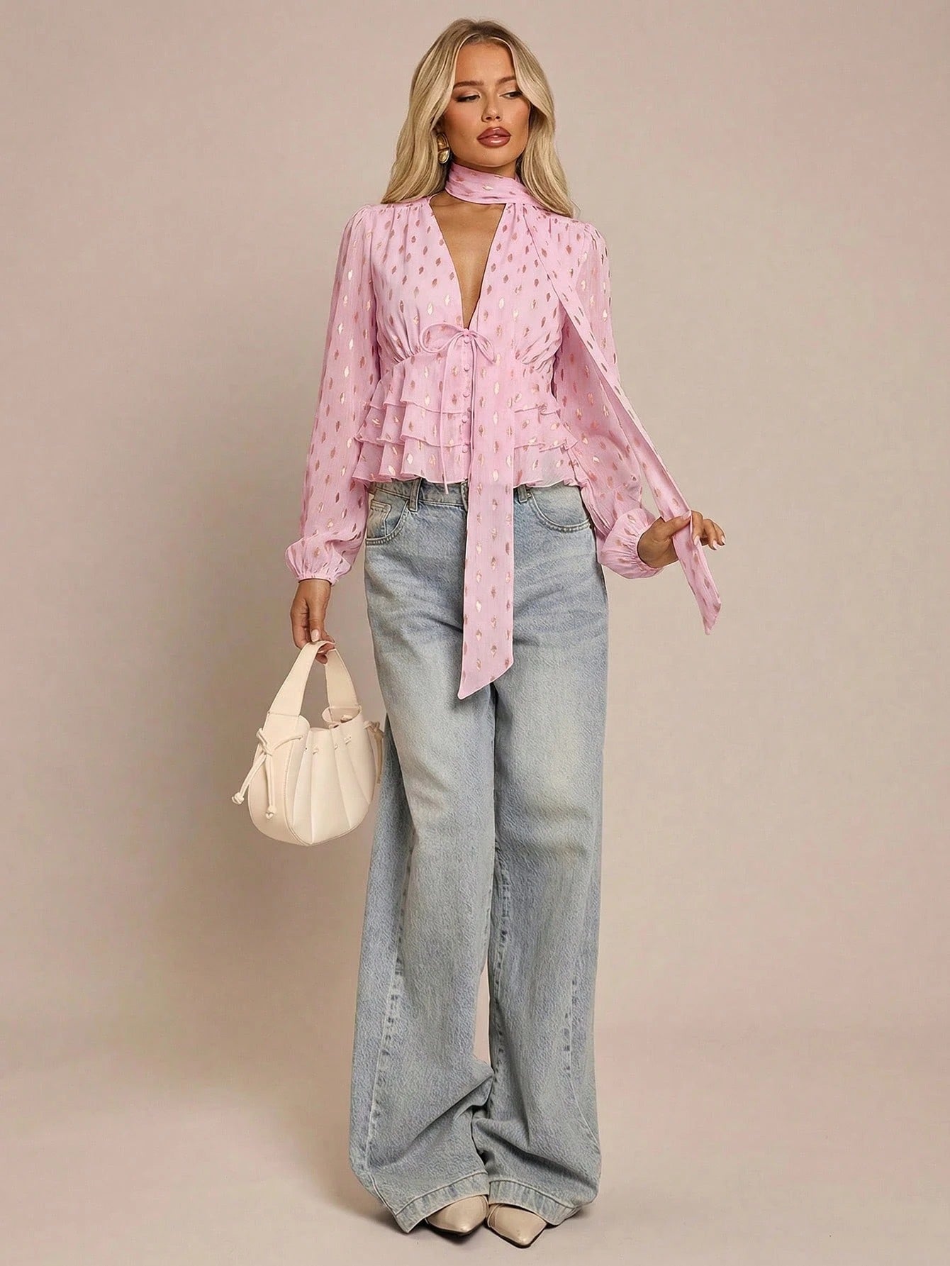 Women's pink long-sleeve chiffon blouse with metallic foil dots, a V-neck, a scarf tie, and a tiered ruffled hem paired with blue jeans.