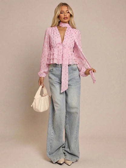Women's pink long-sleeve chiffon blouse with metallic foil dots, a V-neck, a scarf tie, and a tiered ruffled hem paired with blue jeans.