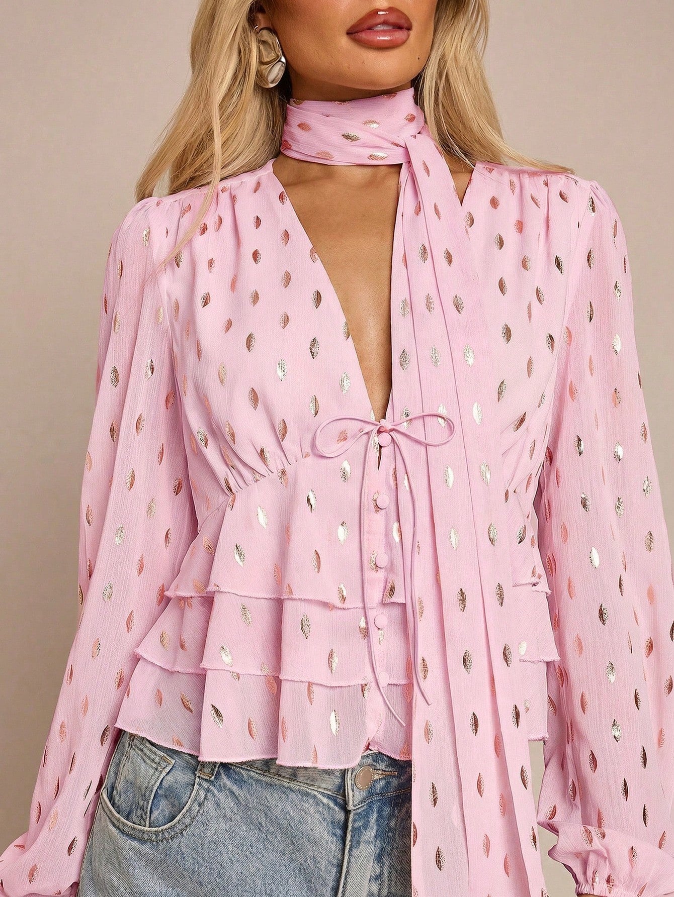 Women's pink long-sleeve chiffon blouse with metallic foil dots, a V-neck, a scarf tie, and a tiered ruffled hem paired with blue jeans.