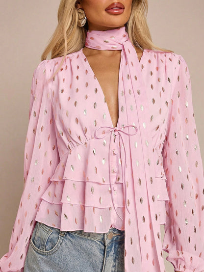 Women's pink long-sleeve chiffon blouse with metallic foil dots, a V-neck, a scarf tie, and a tiered ruffled hem paired with blue jeans.