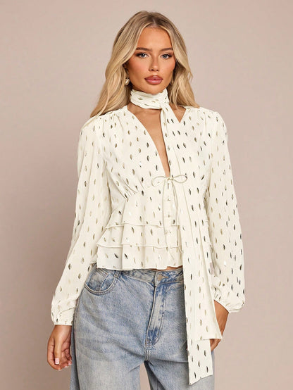 Women's white long-sleeve chiffon blouse with metallic foil dots, a V-neck, a scarf tie, and a tiered ruffled hem paired with blue jeans.
