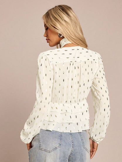 Women's white long-sleeve chiffon blouse with metallic foil dots, a V-neck, a scarf tie, and a tiered ruffled hem paired with blue jeans.