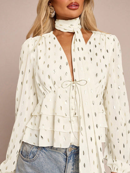 Women's white long-sleeve chiffon blouse with metallic foil dots, a V-neck, a scarf tie, and a tiered ruffled hem paired with blue jeans.