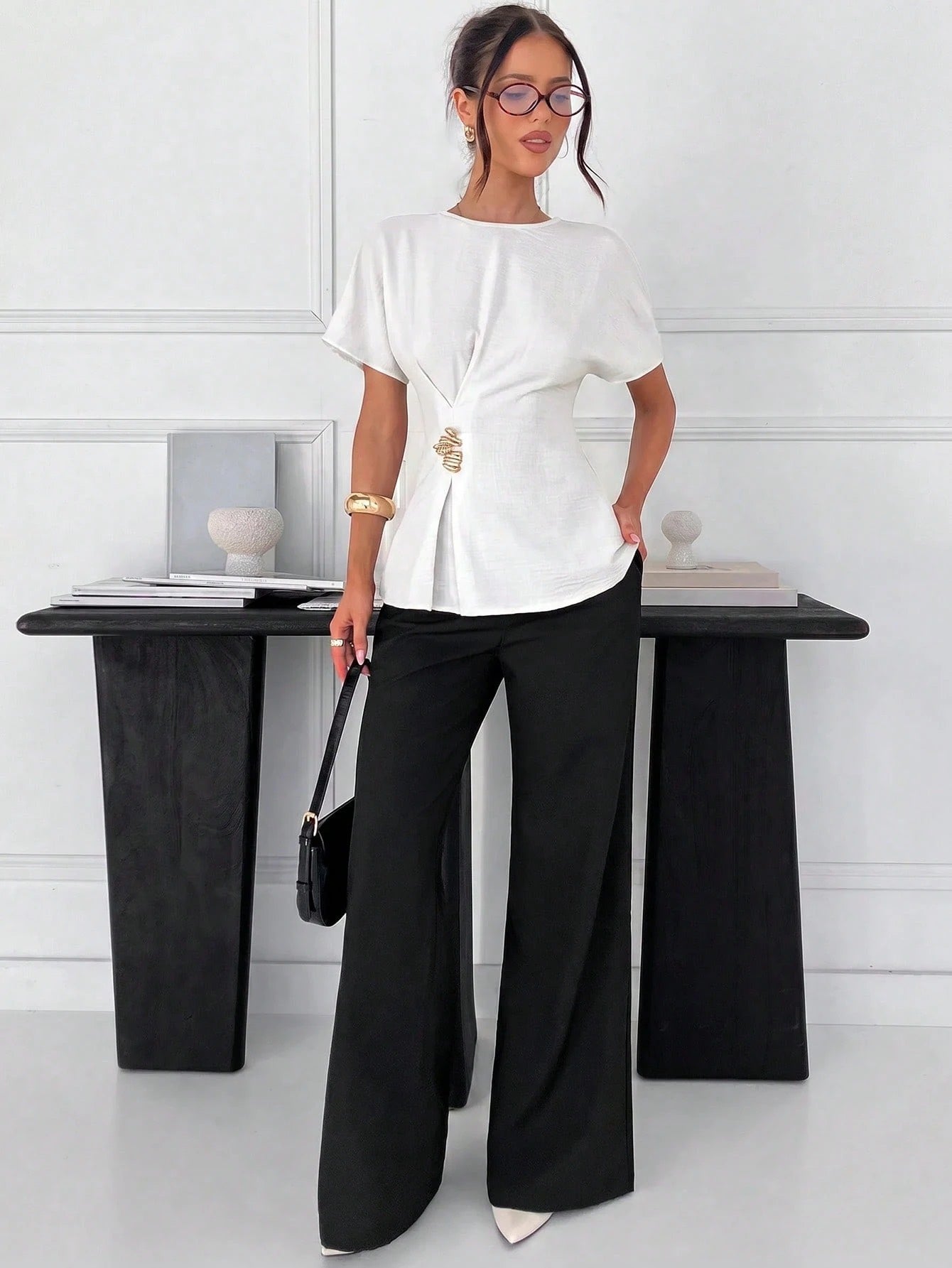 Women's white short-sleeve round neck blouse with a statement gold side waist buckle paired with black trousers for an office look.2