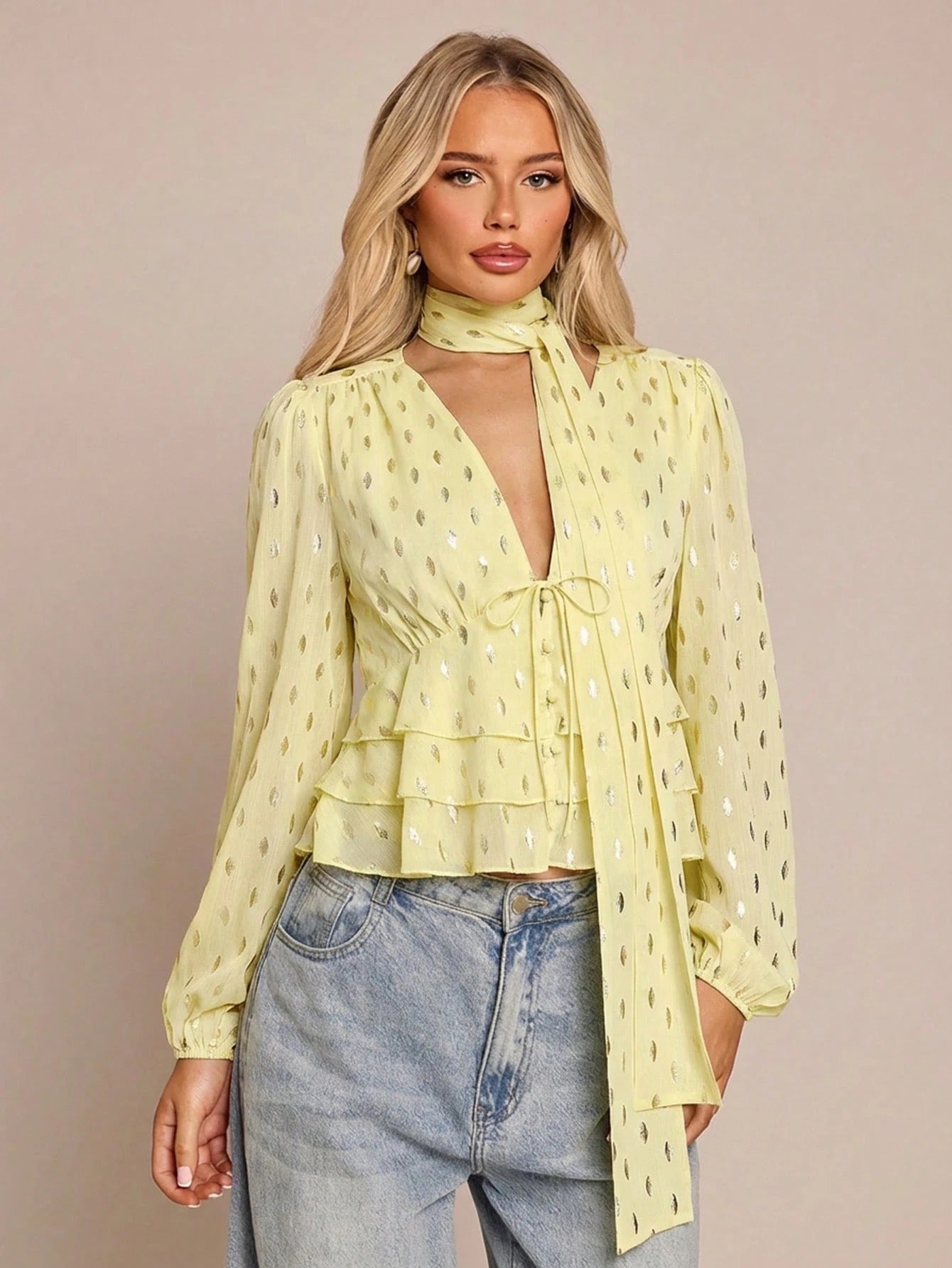 Women's yellow long-sleeve chiffon blouse with metallic foil dots, a V-neck, a scarf tie, and a tiered ruffled hem paired with blue jeans.