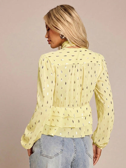 Women's yellow long-sleeve chiffon blouse with metallic foil dots, a V-neck, a scarf tie, and a tiered ruffled hem paired with blue jeans.