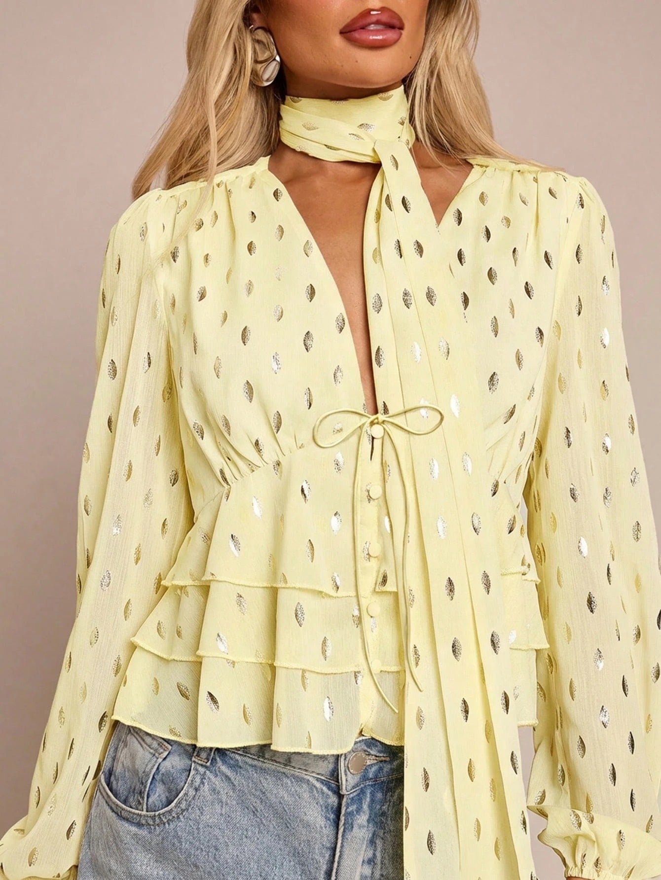 Women's yellow long-sleeve chiffon blouse with metallic foil dots, a V-neck, a scarf tie, and a tiered ruffled hem paired with blue jeans.