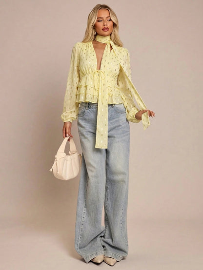 Women's yellow long-sleeve chiffon blouse with metallic foil dots, a V-neck, a scarf tie, and a tiered ruffled hem paired with blue jeans.